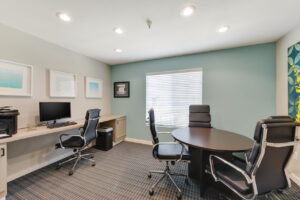 community office room with table and office chairs, desk with computer and large bright window with light blue walls