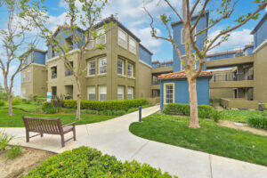 Exterior Overlook at Anaheim Hills, beige and blue residential buildings, meticulous landscaping, bench along sidewalk, lush foliage, photo taken on a sunny day.