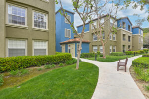 Exterior Overlook at Anaheim Hills, beige and blue residential buildings, meticulous landscaping, bench along sidewalk, lush foliage, photo taken on a sunny day.