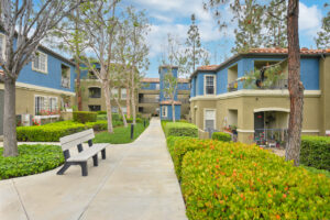 Exterior Overlook at Anaheim Hills, beige and blue residential buildings, meticulous landscaping, bench along sidewalk, lush foliage, photo taken on a sunny day.