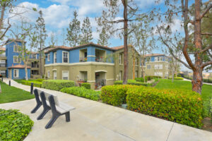 Exterior Overlook at Anaheim Hills, beige and blue residential buildings, meticulous landscaping, bench along sidewalk, lush foliage, photo taken on a sunny day.