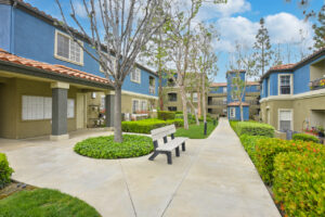 Exterior Overlook at Anaheim Hills, beige and blue residential buildings, meticulous landscaping, bench along sidewalk, lush foliage, photo taken on a sunny day.