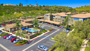 aerial exterior of Overlook at anaheim hills, community center with pool in the back, parking lot with covered spaces, beige and blue residential buildings, sprawling hills in the background, meticulous landscaping, lush foliage, photo taken on a sunny day.