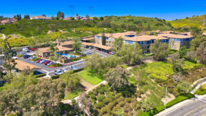 aerial exterior of Overlook at anaheim hills, community center with pool in the back, parking lot with covered parking spaces, beige and blue residential buildings, sprawling hills in the background, lush foliage, meticulous landscaping, photo taken on a sunny day.