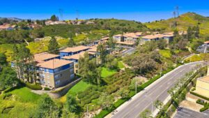 Aerial Exterior of The Overlook at Anaheim Hills, beige and blue residential buildings, meticulous landscaping, lush foliage, sprawling hills in the background, photo taken on a sunny day.