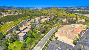 aerial exterior of the Overlook at Anaheim Hills, meticulous landscaping, lush foliage, view of surrounding businesses, beige and blue residential buildings, sprawling hills in the background, surrounding neighborhoods in the distance, photo taken on a sunny day.