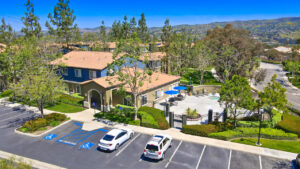 aerial exterior of community center, parking in front, meticulous landscaping, outdoor patio with pergola grilling area and hot tub, sprawling hills in the distance, lush foliage, photo taken on a sunny day.