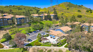 aerial exterior of the Overlook at Anaheim hills, community center with outdoor patio including pergola grilling area, hot tub, patio furniture, residential buildings adjacent to community center, sprawling hills in the background, meticulous landscaping, lush foliage, photo taken on a sunny day.