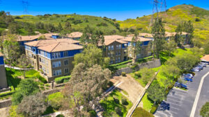 aerial exterior of the Overlook at Anaheim Hills, lush foliage, meticulous landscaping, parking adjacent to buildings, sprawling hill behind residential buildings, photo taken on a sunny day.