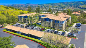 Aerial Exterior of The Overlook at Anaheim Hills, parking lot with covered parking spaces, beige and blue residential buildings, meticulous landscaping, neighborhoods and sprawling hills in background, lush foliage, photo taken on a sunny day.