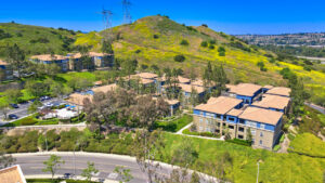 aerial exterior of the Overlook at Anaheim hills, meticulous landscaping, lush foliage, beige and blue residential buildings, sprawling hill in the background, parking along residential buildings, photo taken on a sunny day.