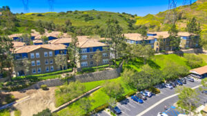 Aerial Exterior of The Overlook at Anaheim Hills, parking along residential buildings, beige and blue residential buildings, sprawling hills in the background, meticulous landscaping, photo taken on a sunny day.